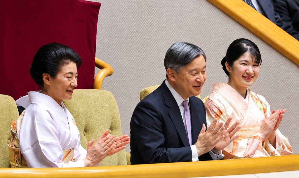 Three people sit in a formal arena, clapping: two women in kimonos flanking a man in a suit, smiling.