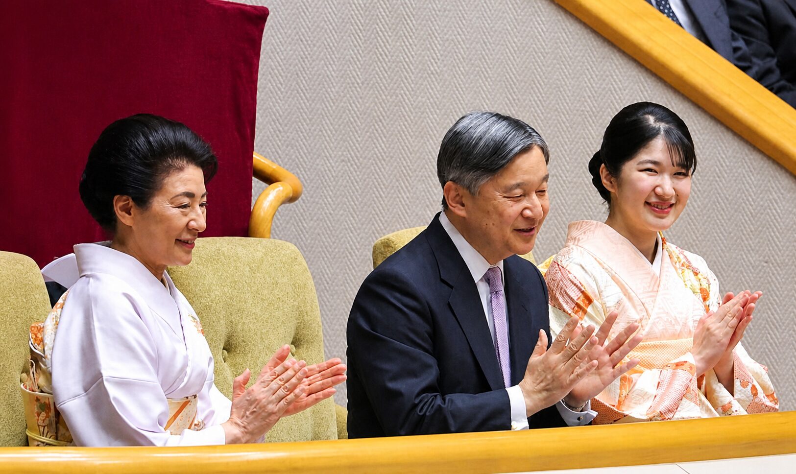 Three people sit in a formal arena, clapping: two women in kimonos flanking a man in a suit, smiling.