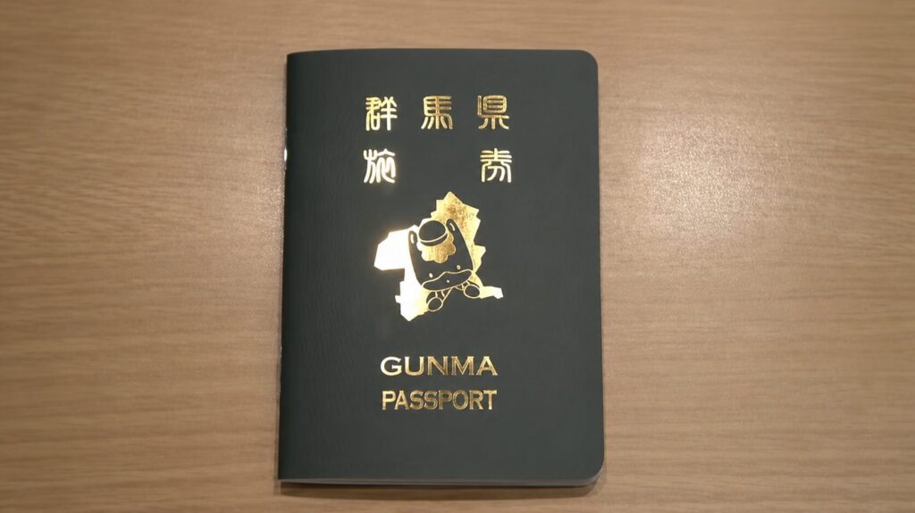 Dark green passport with gold embossed text and mascot; reads 'GUNMA PASSPORT' on a wooden surface.