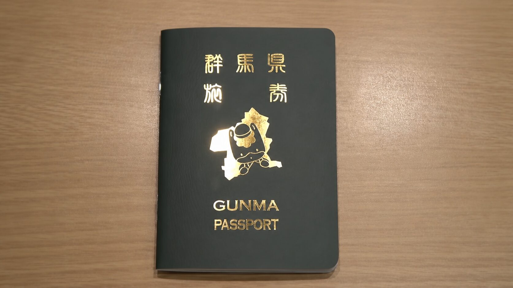 Dark green passport with gold embossed text and mascot; reads 'GUNMA PASSPORT' on a wooden surface.