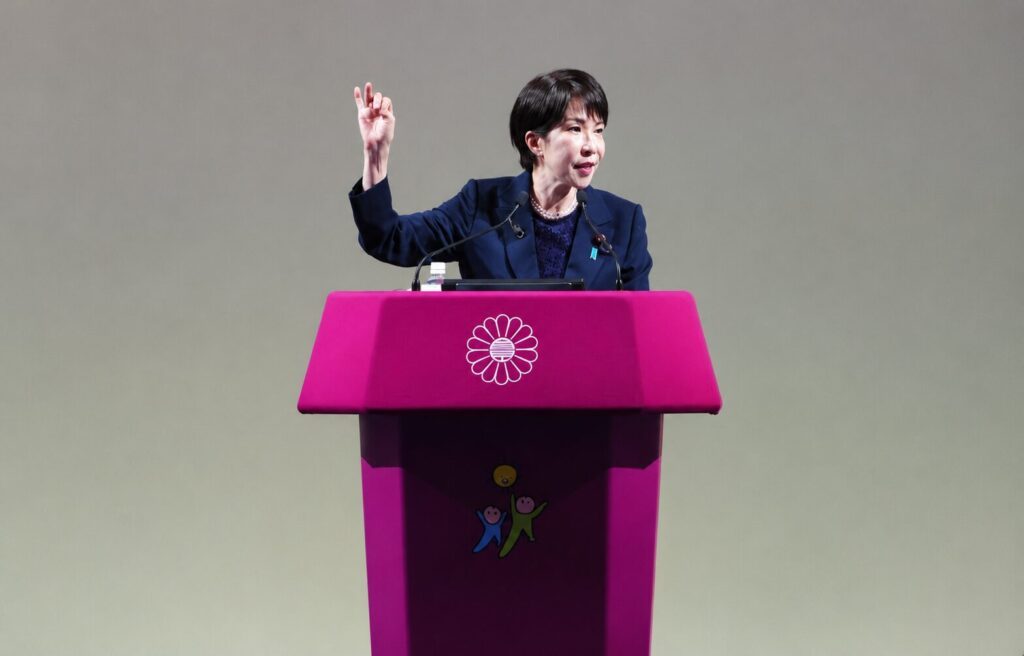 Female speaker at a bright pink podium with a white flower emblem, delivering a speech with microphones on stage.