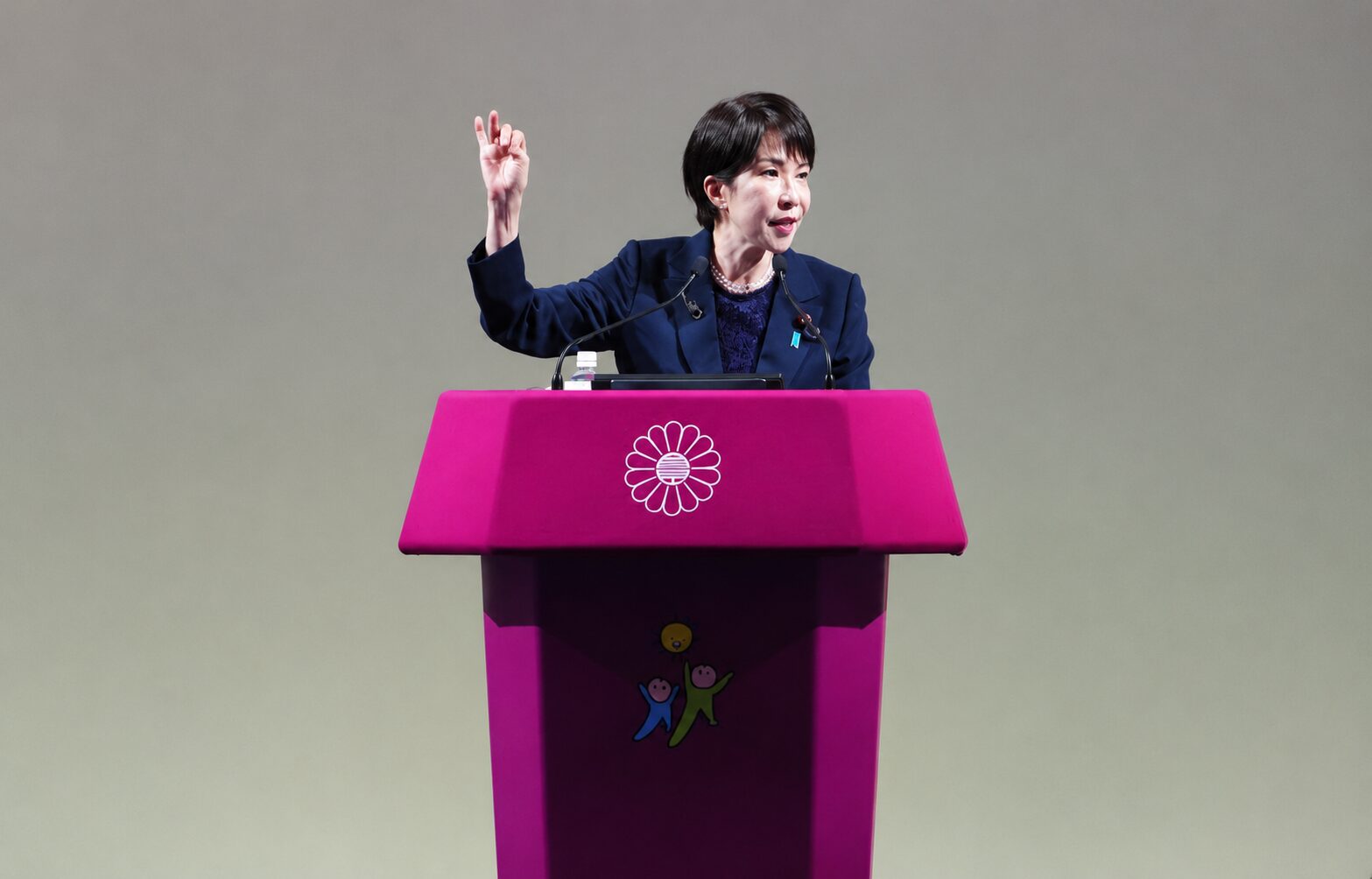 Female speaker at a bright pink podium with a white flower emblem, delivering a speech with microphones on stage.