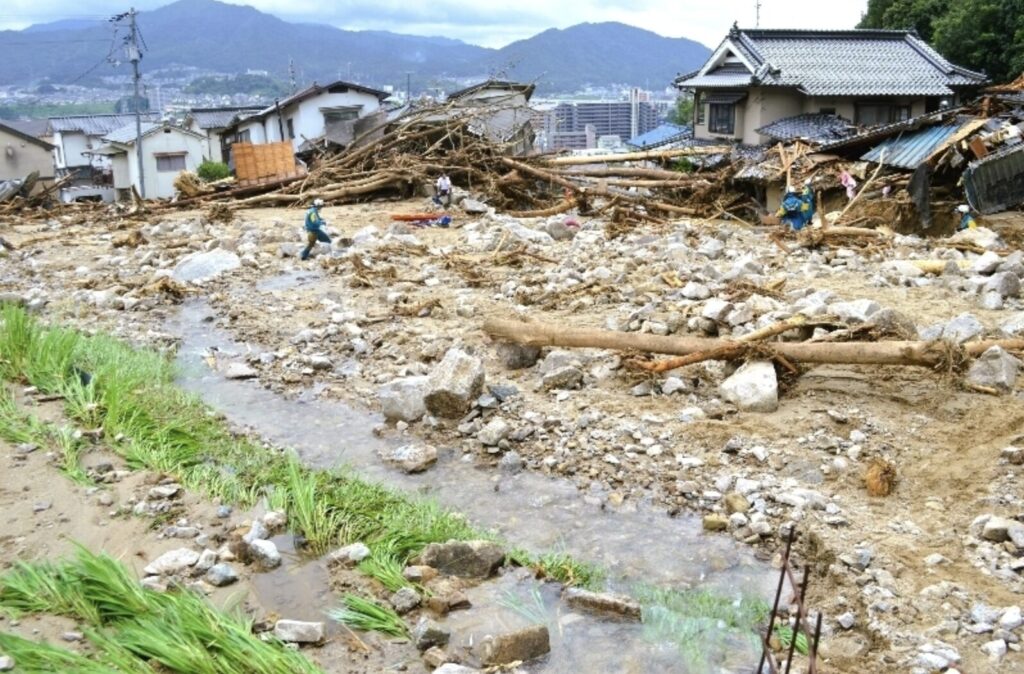 Debris and collapsed houses after a landslide or flood; workers in gear survey rubble and mud in a ruined street with mountains in the background