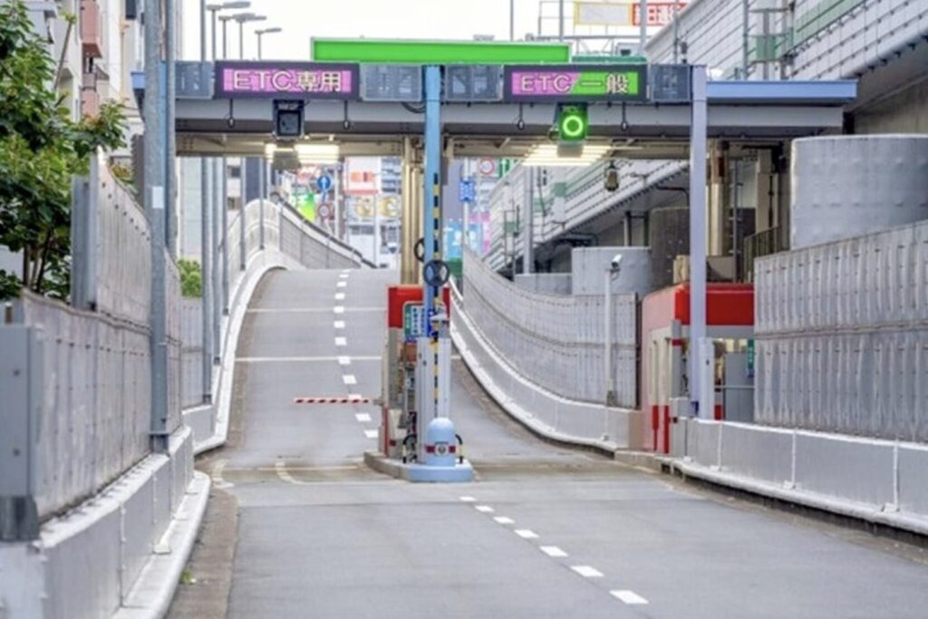 Entrance to a sunken city road with a curved ramp, concrete barriers, and metal fencing; overhead signage visible in urban setting.