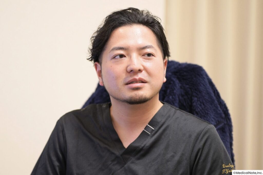 Man with short dark hair in black scrubs, seated indoors, looking to the right with a neutral expression against a beige wall and dark chair behind him.