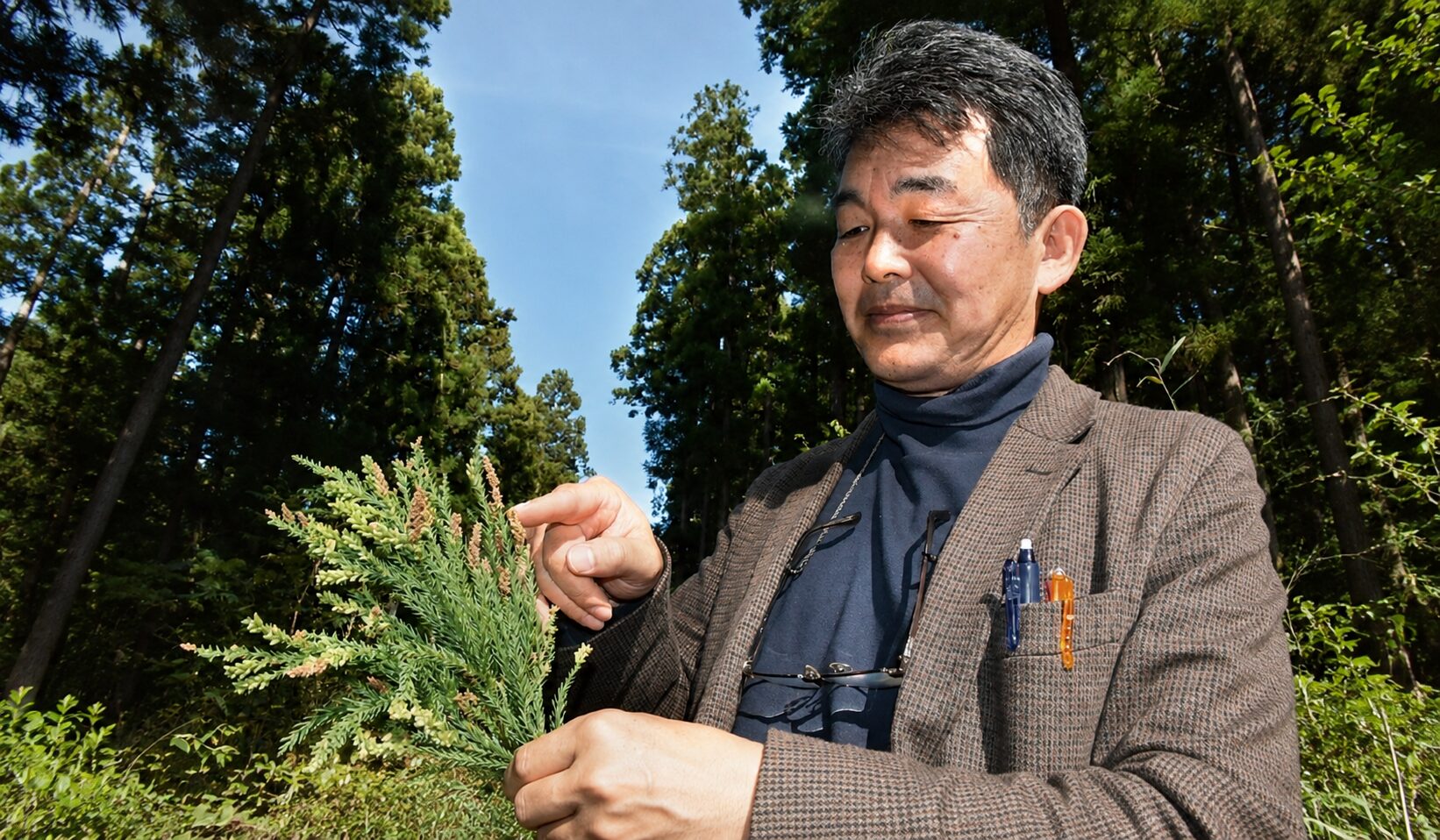 Man in a brown blazer and dark turtleneck outside in a forest, closely examining a small evergreen branch with needle-like leaves and tiny cones.