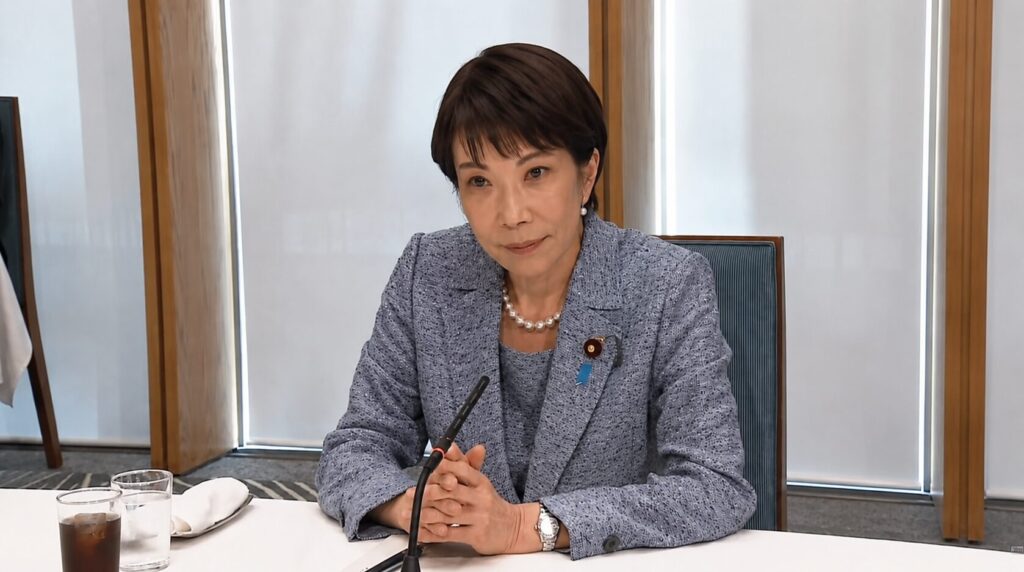 Professional woman in a blue textured blazer sits at a conference table with a microphone, clasped hands, pearls, and a lapel pin.