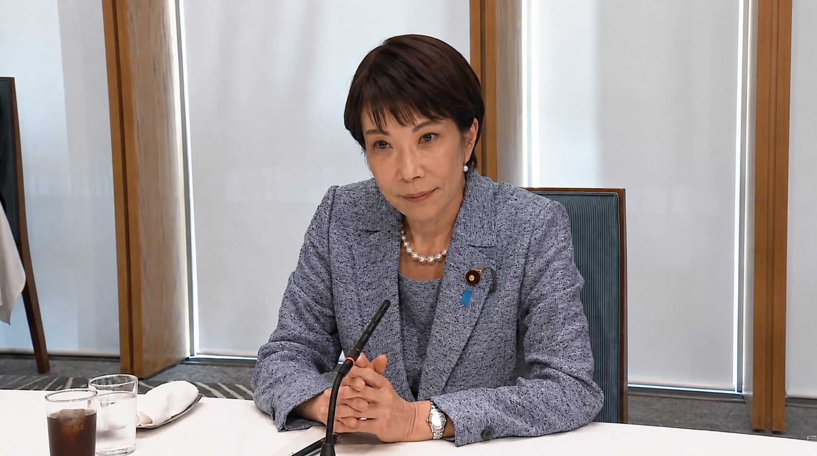 Professional woman in a blue textured blazer sits at a conference table with a microphone, clasped hands, pearls, and a lapel pin.