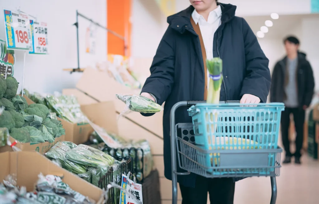 Shopper in a dark jacket pushes a cart while selecting bok choy from the produce section in a grocery store.