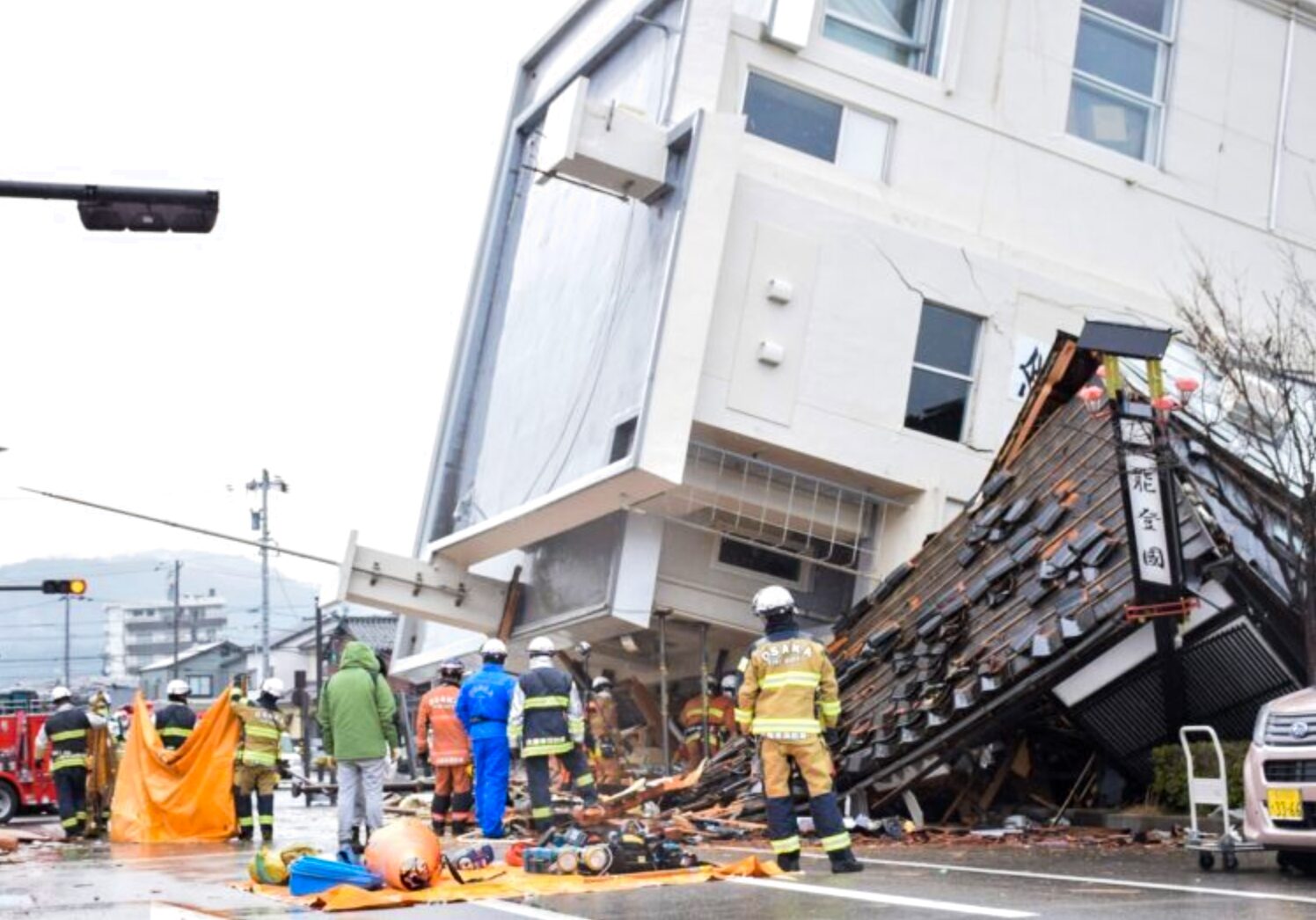 Firefighters and responders survey a collapsed building with debris and equipment scattered on a wet street.